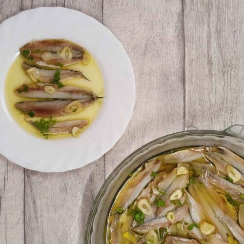 A white plate with the marinated fish on it, covered with olive oil and decorated with parsley. At the bottom of the photo there is a large glass dish with all the marinated fish in it.