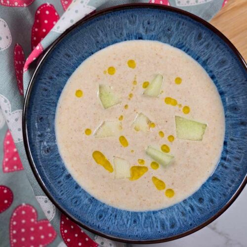 A blue bowl with the white soup in it, with yellow olive oil spots on top and green pieces of melon in it